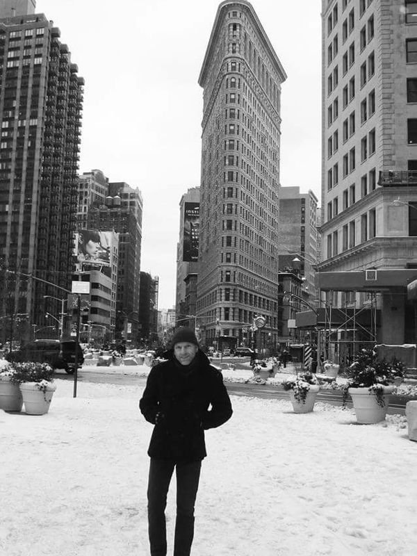 A black and white photo of a man standing in front of the Flatiron building in New York City, snow on the ground