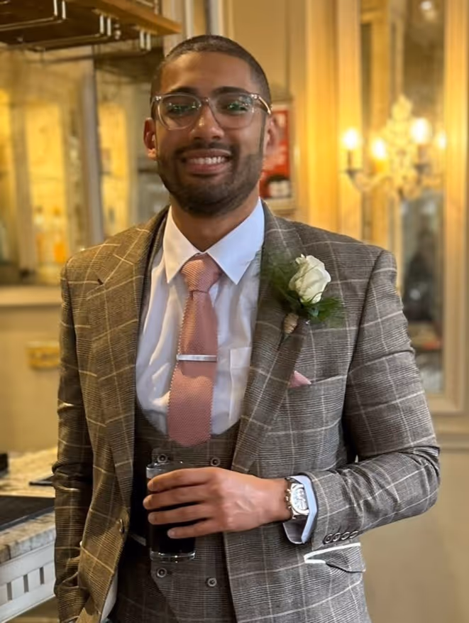 Smiling younger man in a check suit and pink tie holds a drink at an indoor event.