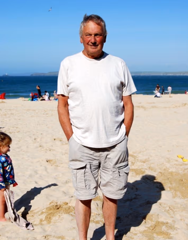 Older man standing on sandy beach wearing white t-shirt and light shorts with ocean in background.