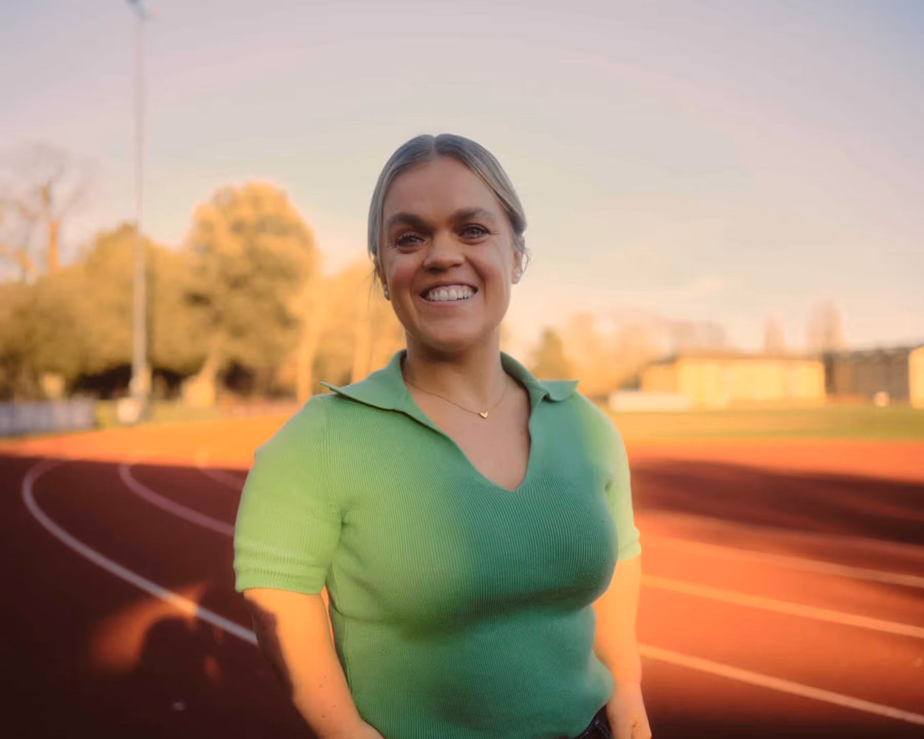 Ellie Simmonds smiling and posing in a green polo shirt at a sunny athletics track.