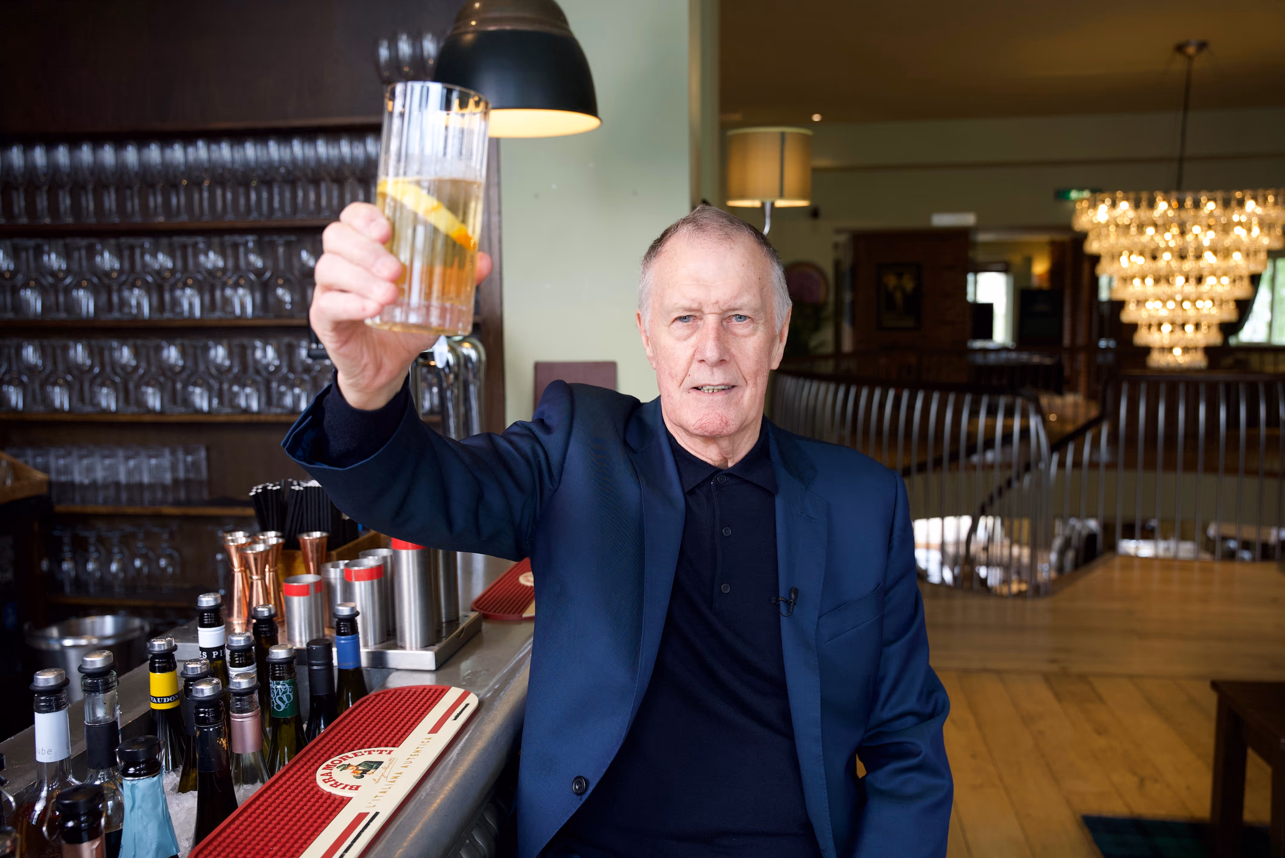 Geoff Hurst wearing a dark navy suit jacket, raising a drink at a bar.