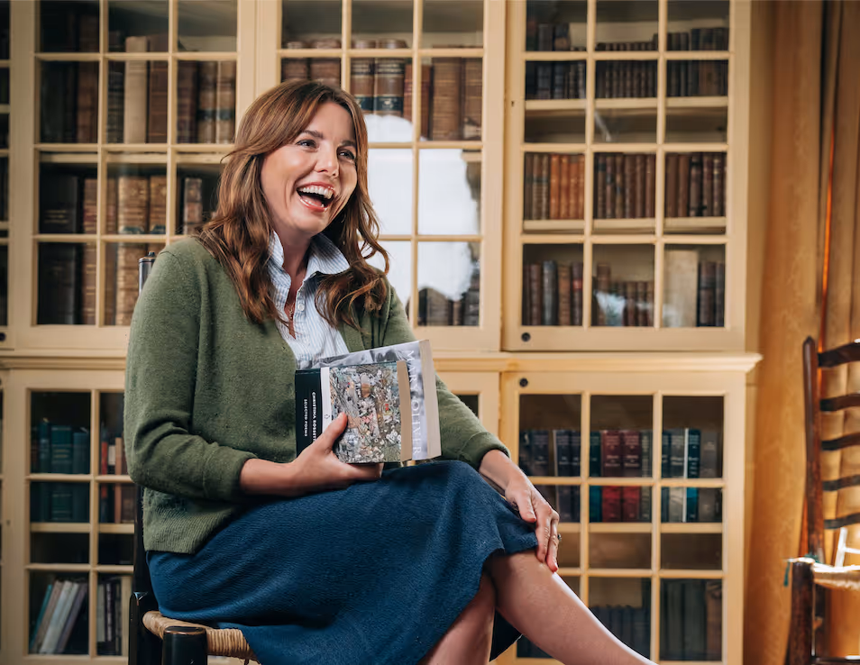 Ophelia Lovibond laughing, holding two books in front of a large white bookcase.