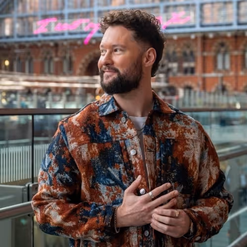 Calum Scott wearing a patterned jacket, looking to the side from a glass balcony.