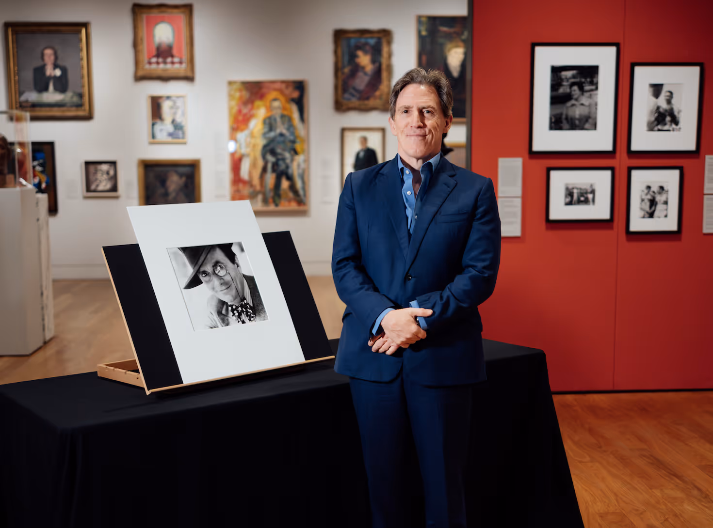Rob Brydon posing next to a black and white portrait of Barry Humphries inside a picture gallery. 