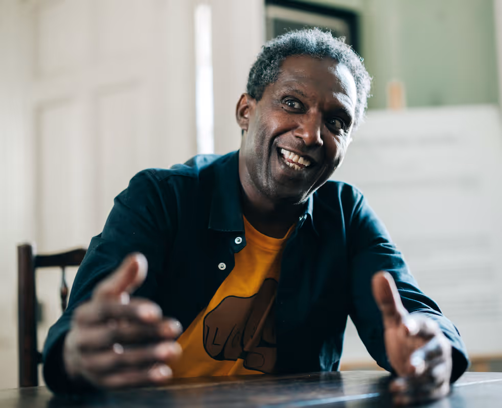 Lemn Sissay smiling and sitting at a wooden table and desk.