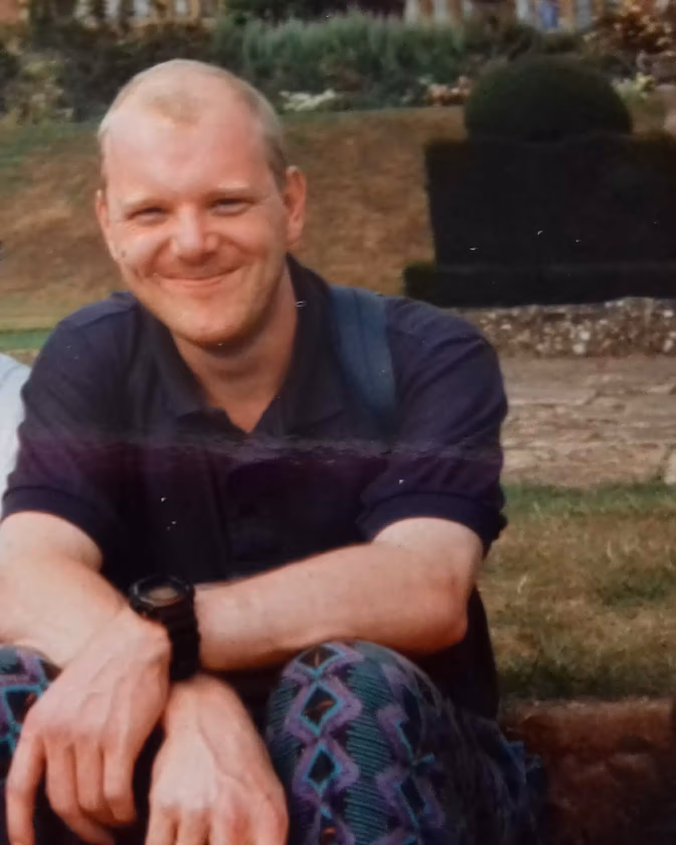 Smiling man with short hair wearing a dark shirt and patterned trousers sitting outdoors on grass.