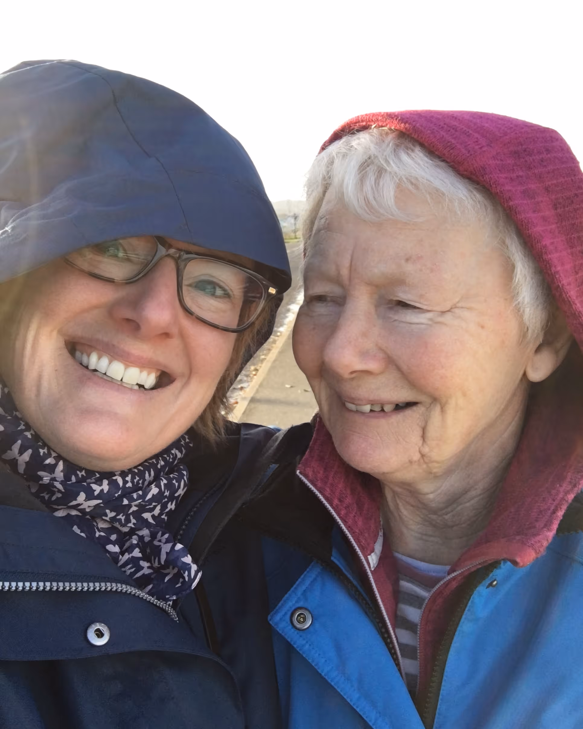 One younger and one elderly woman wearing hooded jackets smiling closely outdoors on a sunny day.