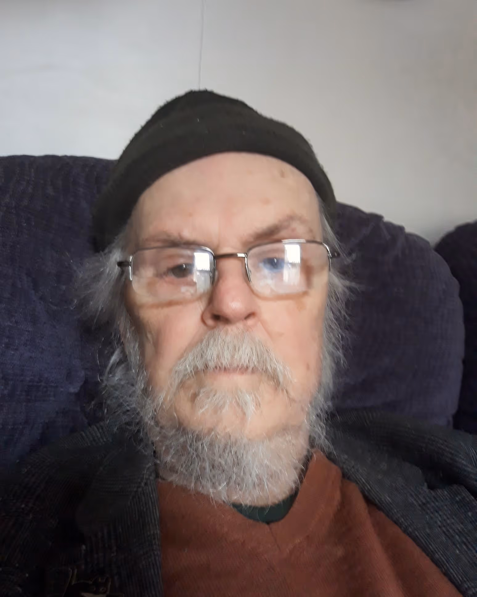 Elderly man with grey beard, glasses, black beanie hat and dark jacket sitting on a purple chair.