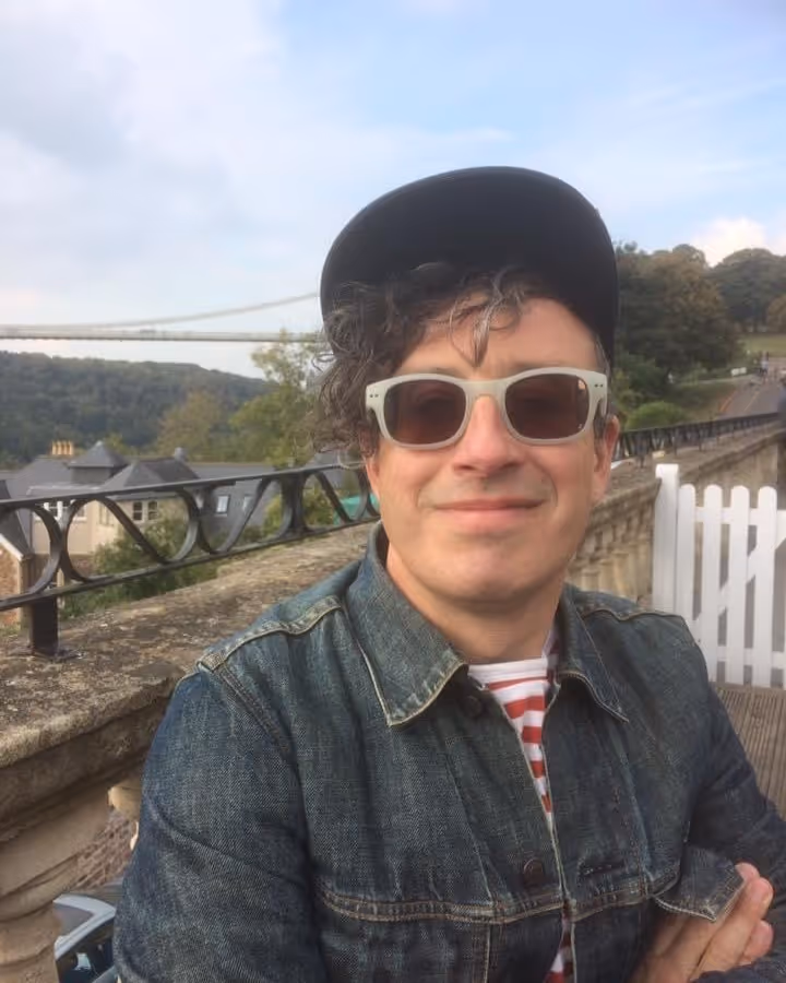 Man wearing sunglasses, a black cap, and a denim jacket, smiling outdoors with a bridge and trees in the background.
