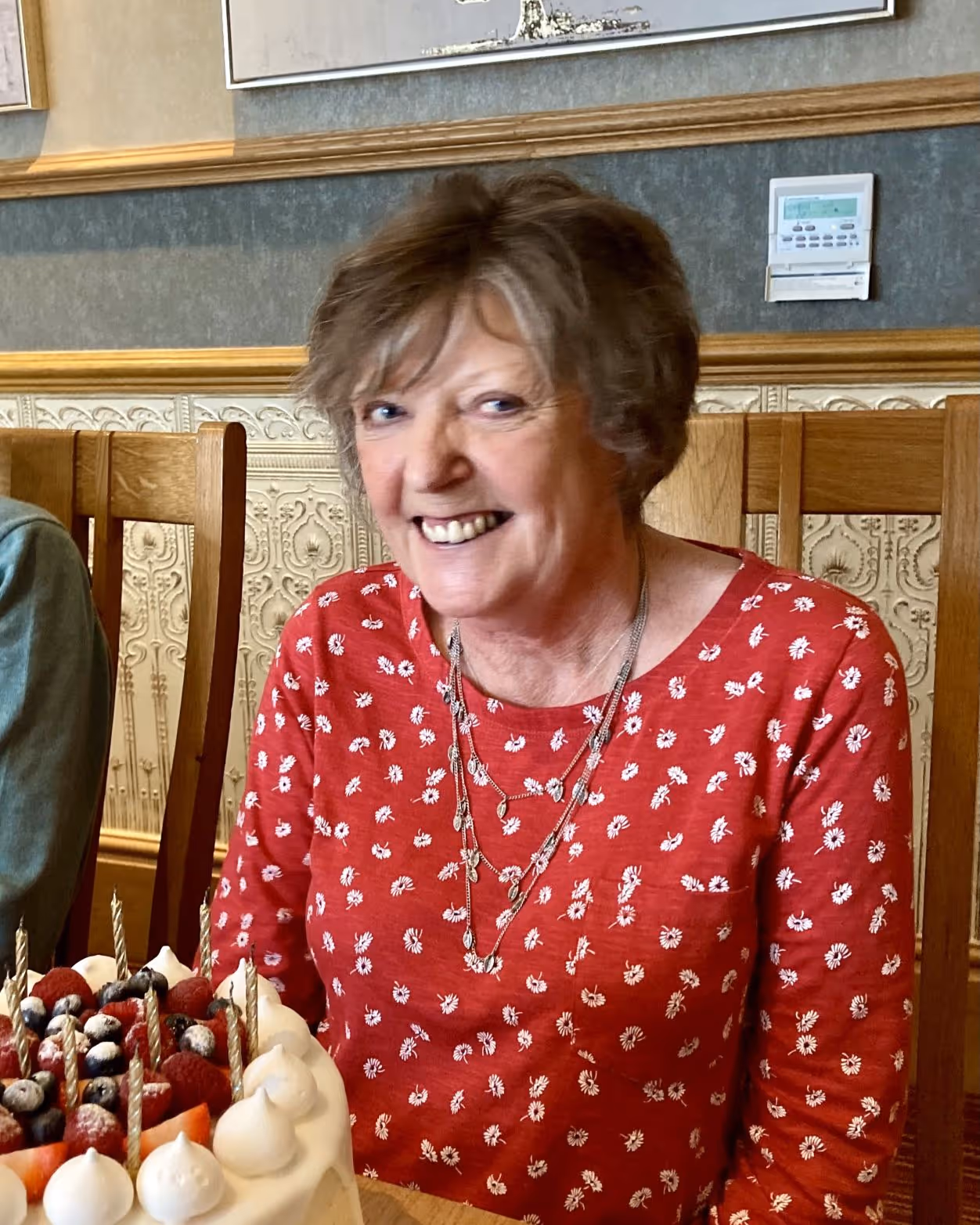 Smiling woman in red floral shirt sitting at a table with a fruit-topped cake with candles.