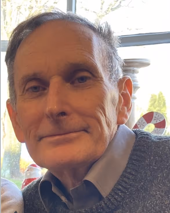 Close-up of an older man with grey hair wearing a collared shirt and grey sweater, indoors by a window.